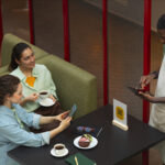 Two women sitting at a café table using their phones to place an order while a waiter records it on a tablet, representing customers booking through aggregator apps.