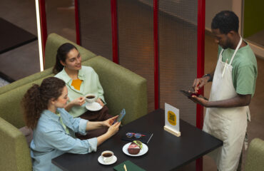 Two women sitting at a café table using their phones to place an order while a waiter records it on a tablet, representing customers booking through aggregator apps.