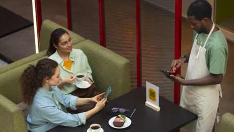 Customer paying through aggregators Two women sitting at a café table using their phones to place an order while a waiter records it on a tablet, representing customers booking through aggregator apps.